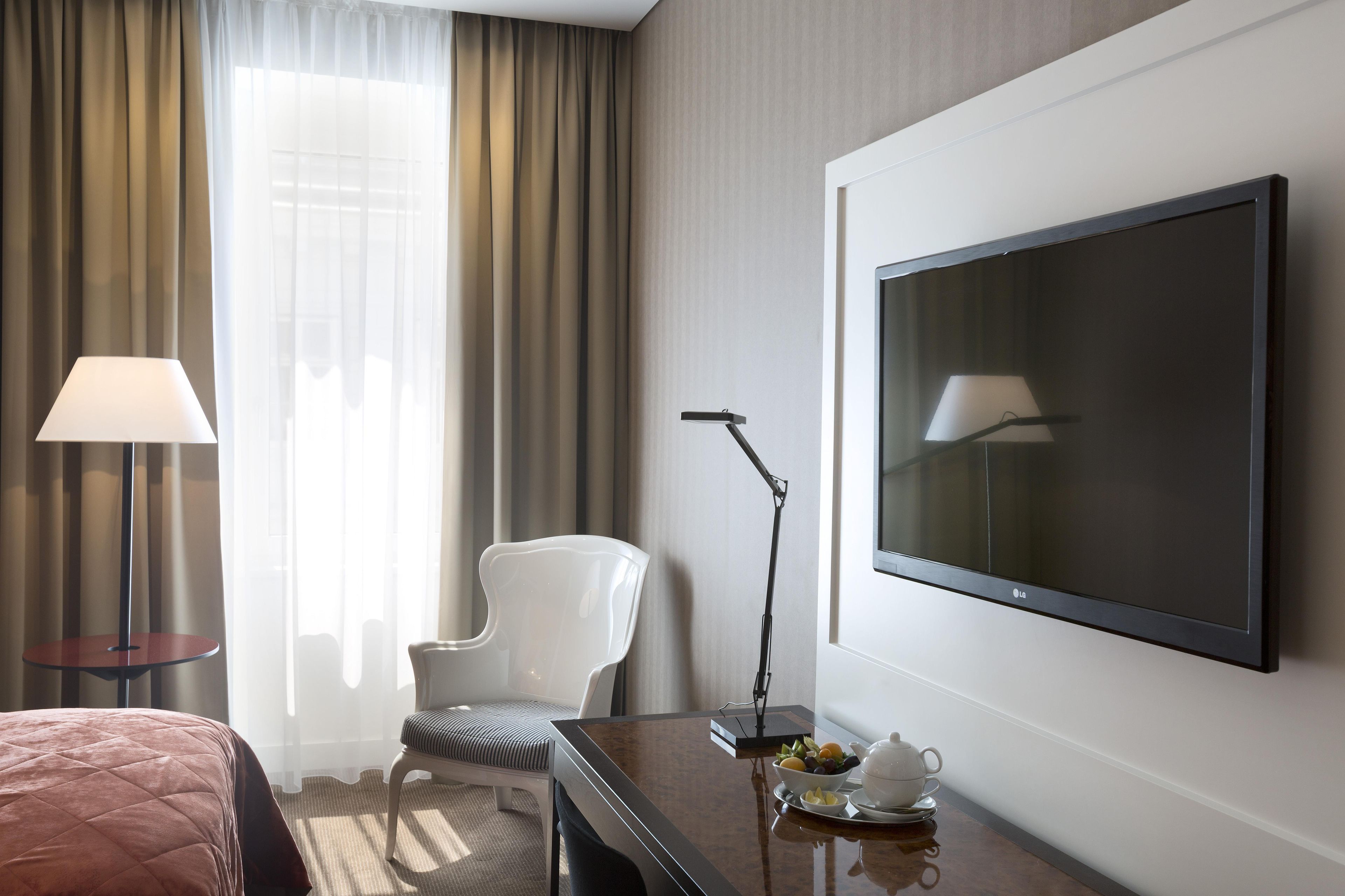 A hotel room with a bed, a desk with a lamp, a chair, a television, and a small table with a teapot and fruit.