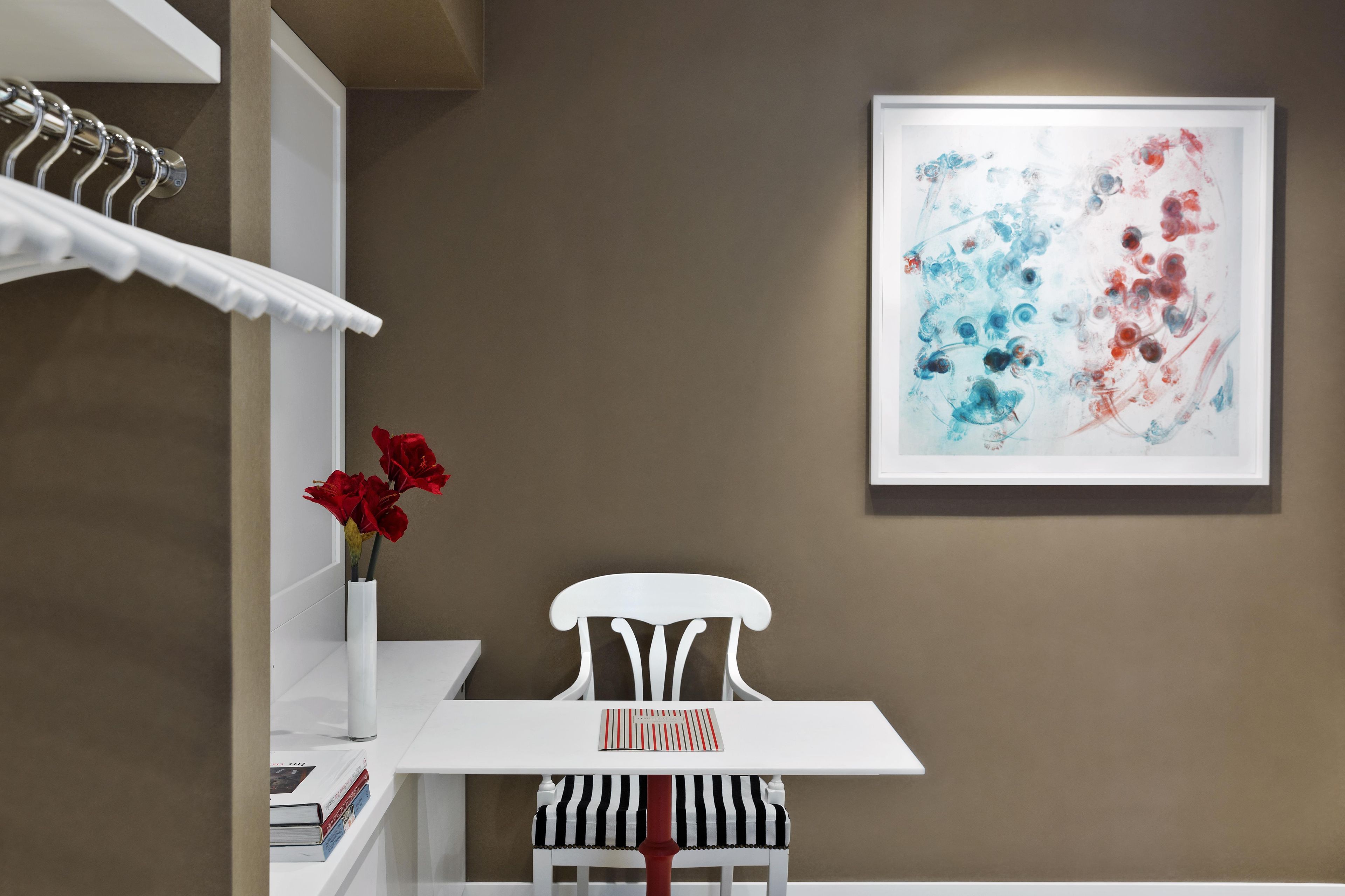Una habitación con una mesa blanca, una silla blanca, un cuadro abstracto enmarcado y un jarrón con flores rojas.