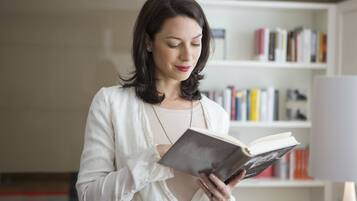 Una persona leyendo un libro en una habitación con una estantería al fondo.