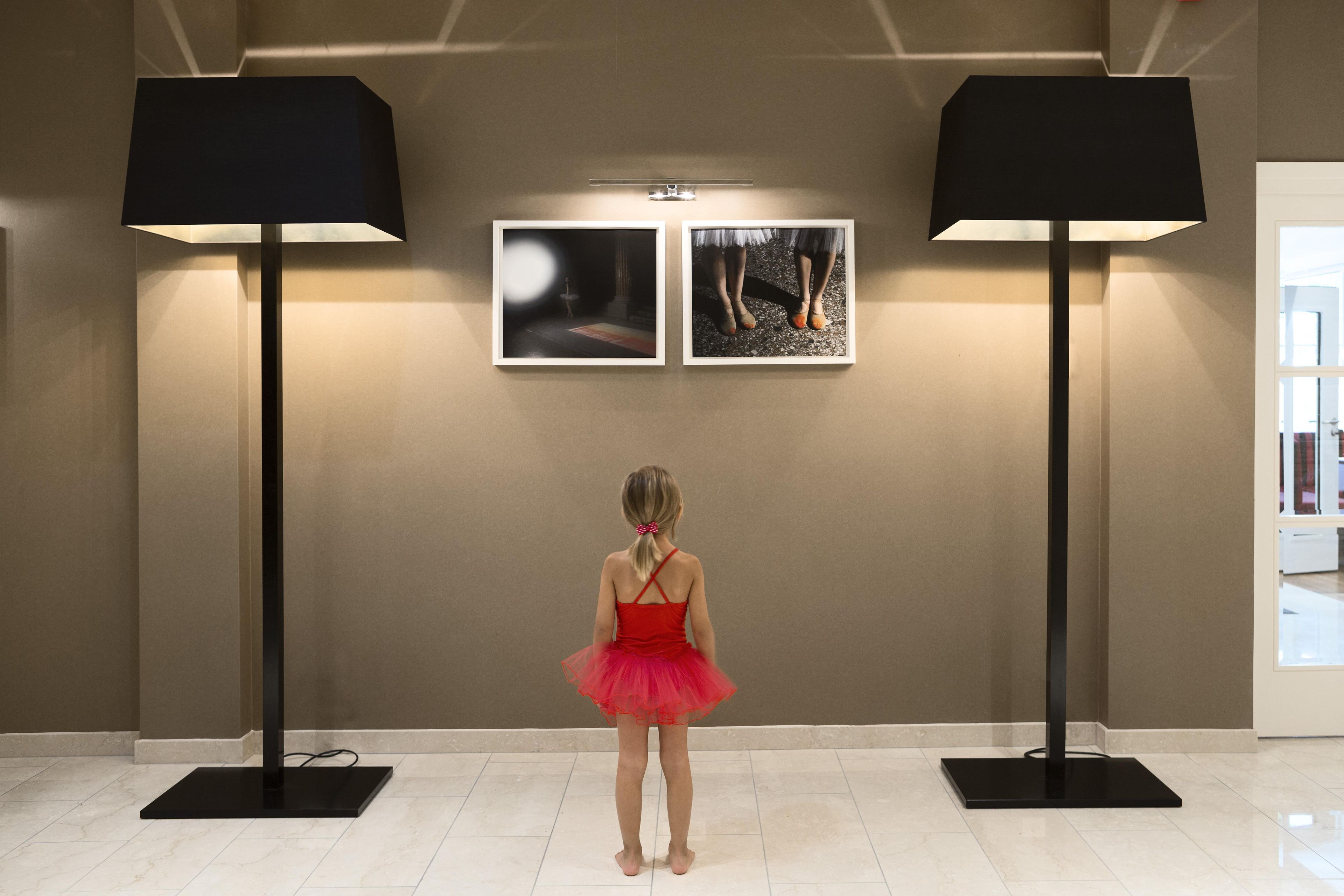 Une jeune fille, vêtue d’une robe rouge, se tient dans un couloir. Deux tableaux encadrés sont accrochés au mur, et de part et d’autre, deux lampadaires sont placés sur le sol.