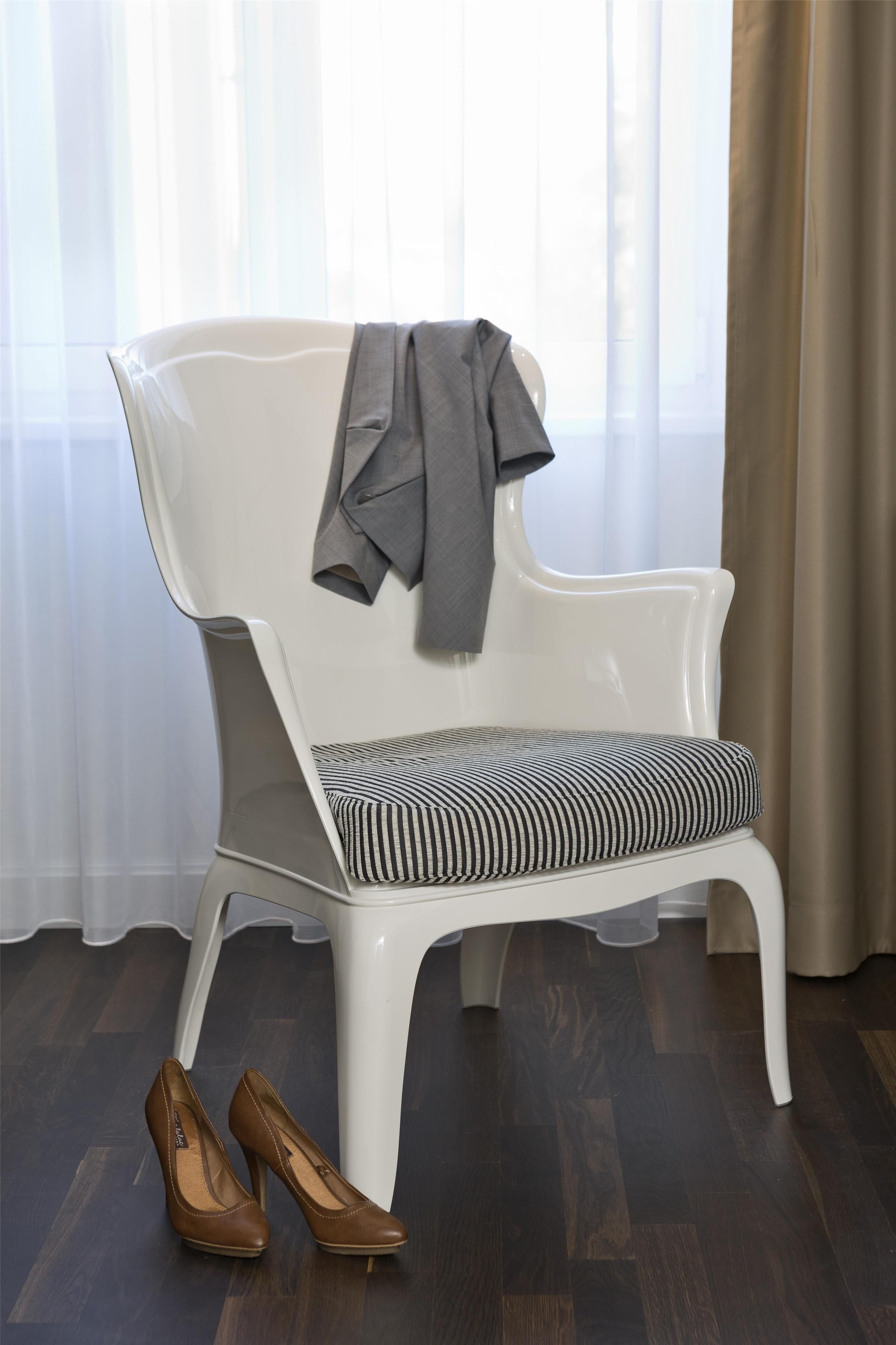 Una silla blanca con un cojín a rayas y una chaqueta gris colgada. Un par de zapatos de tacón marrón está colocado en el suelo junto a la silla.