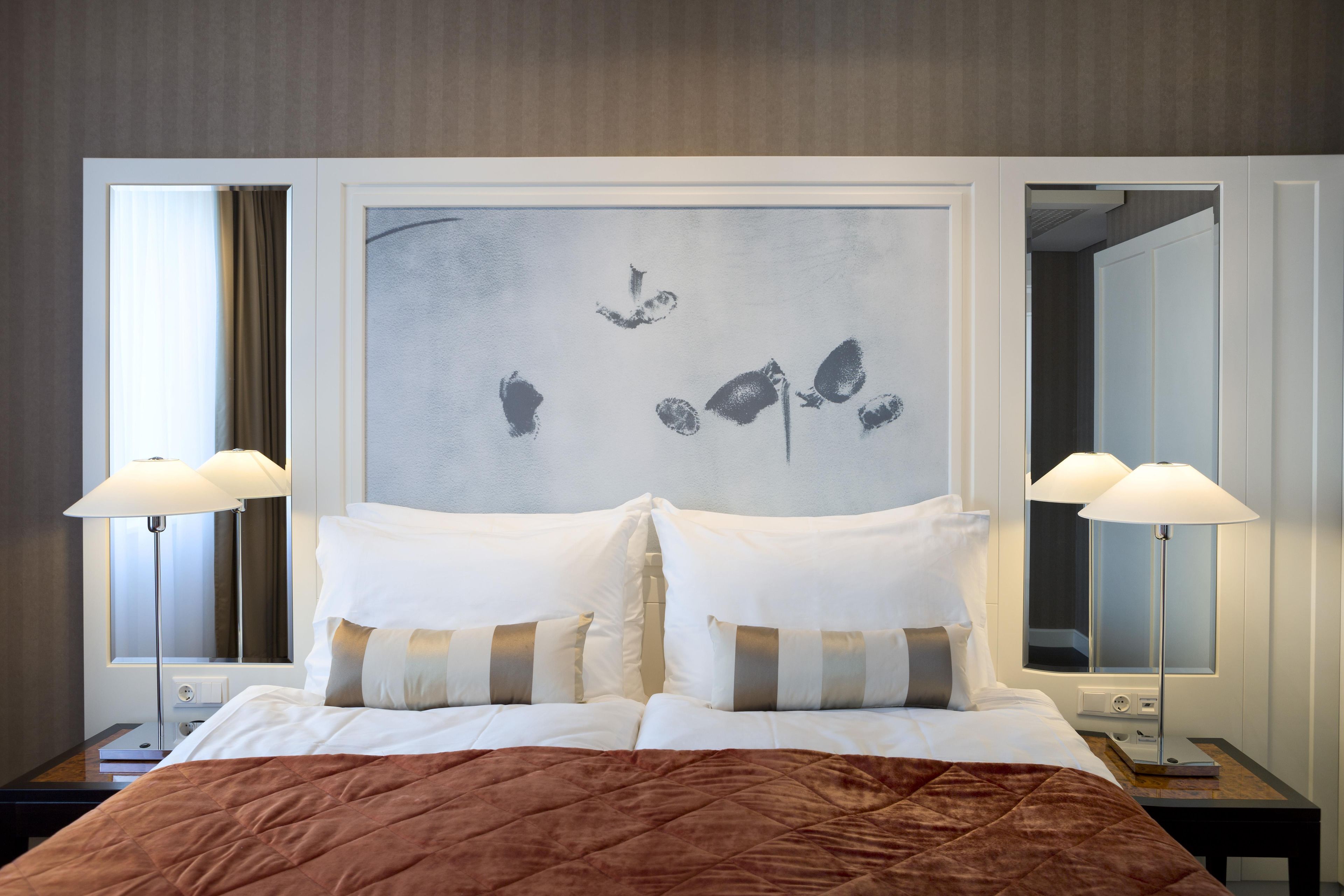 Habitación de hotel con una cama, dos lámparas de noche, un espejo grande y una obra de arte en la pared.