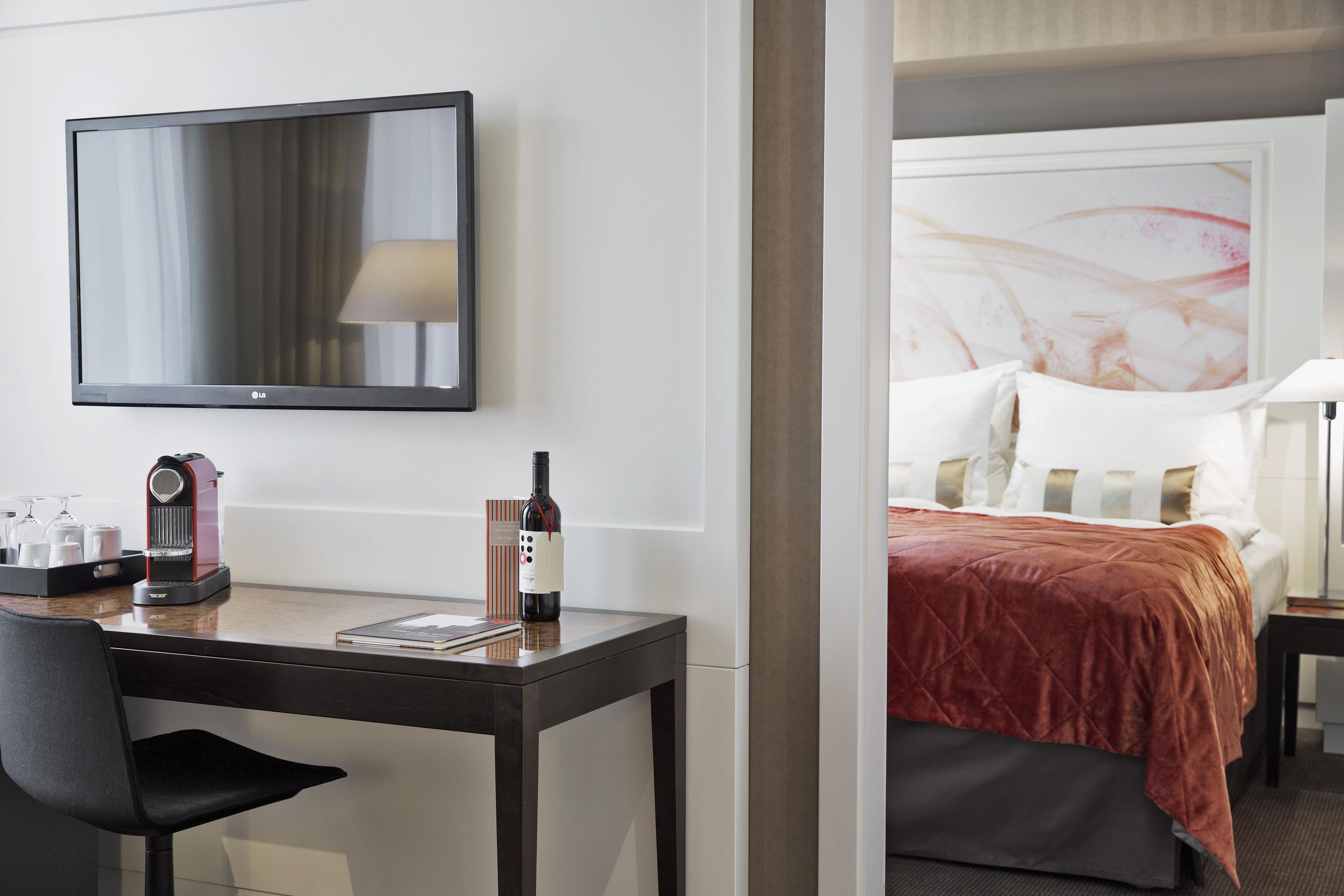 Quarto de hotel com cama, escrivaninha com cafeteira, garrafa de vinho e televisão na parede.