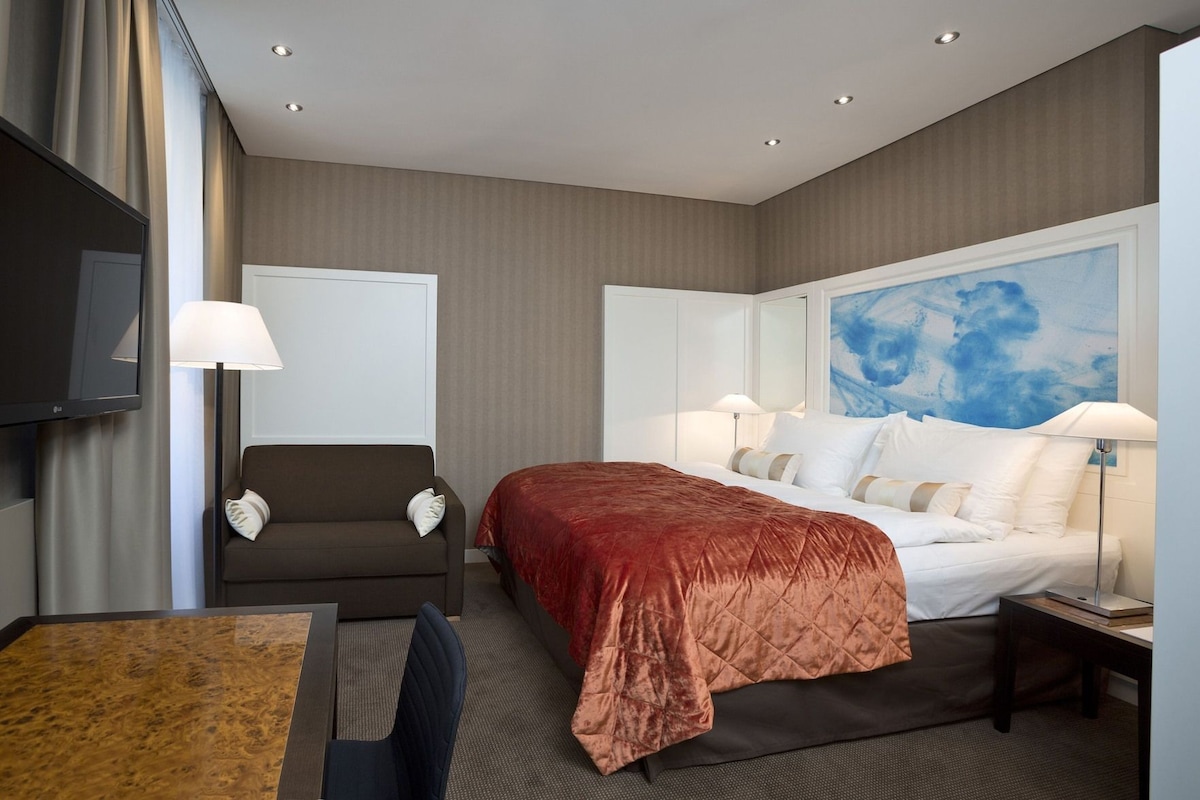 Habitación de hotel con cama, sofá, escritorio, televisor y un cuadro en la pared.