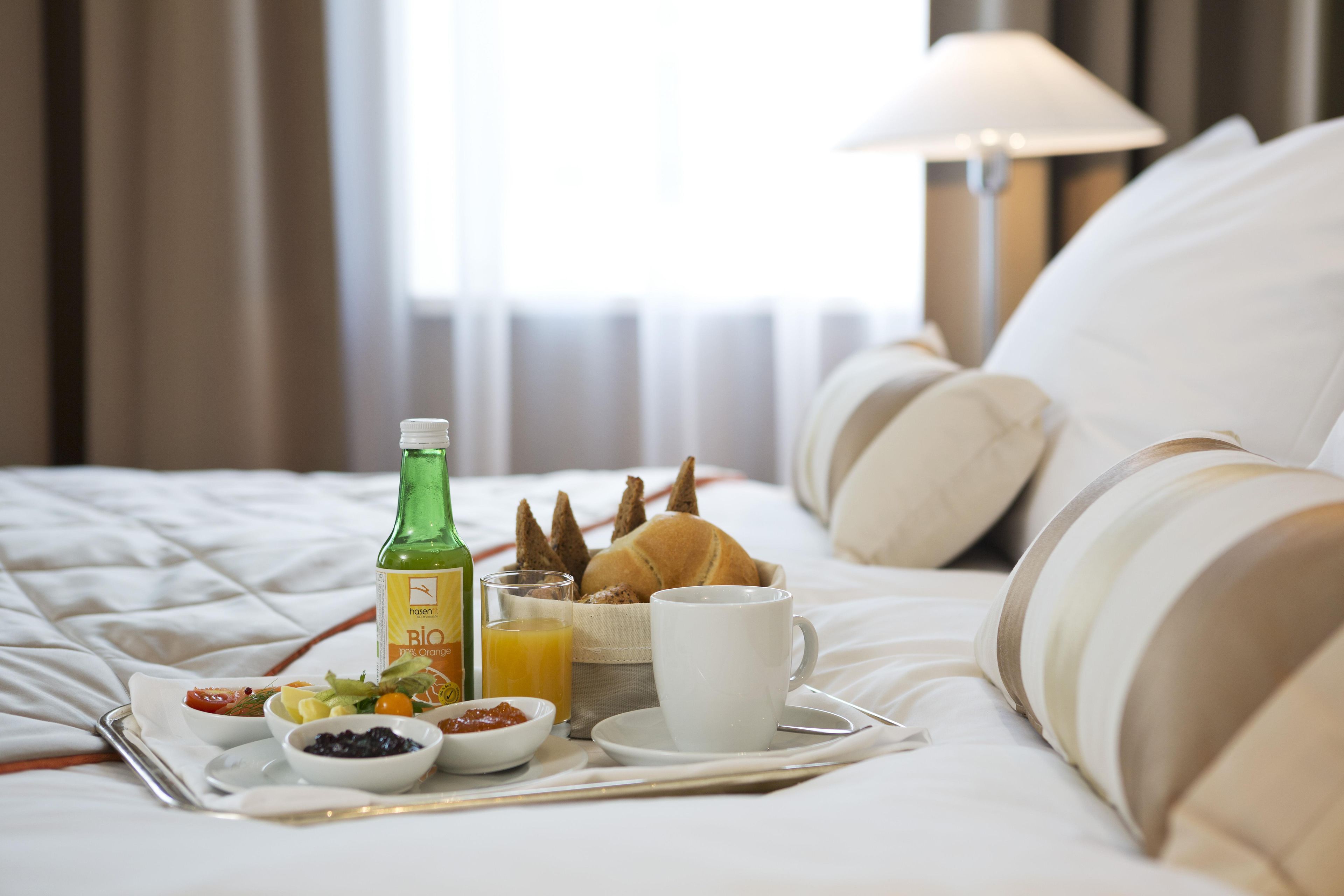 Una bandeja de desayuno bien preparada sobre una cama, con una botella de jugo de naranja, croissants, y tazones de fruta y mermelada.