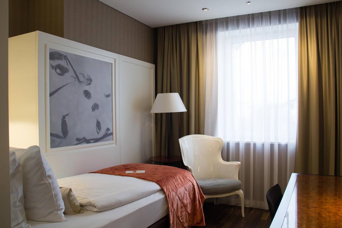 A hotel room with a bed, a chair, a desk, a lamp, and a large abstract painting on the wall.