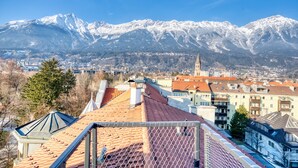 Un paisaje urbano con montañas nevadas al fondo, una aguja de iglesia y edificios residenciales con techos de tejas rojas.