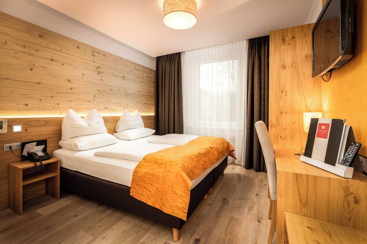 A hotel room with a wooden headboard, a bed with white linens and an orange throw, a bedside table with a phone and a lamp, a TV mounted on the wall, and a window with curtains.