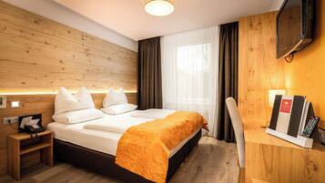 Habitación de hotel con cabecera de madera, cama con sábanas blancas y una manta naranja, mesita de noche con teléfono y lámpara, televisor montado en la pared y ventana con cortinas.
