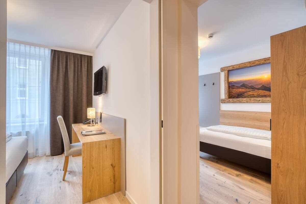 A modern hotel room with a wooden desk, a bed, a TV, and a large window with curtains.
