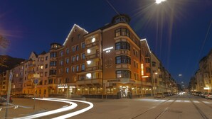 Un hotel de varios pisos con entrada en esquina, letreros iluminados para un restaurante y un bar, y una línea de tranvía que recorre la calle.