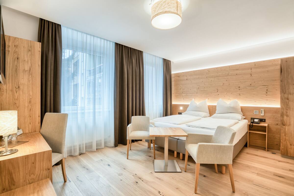 A modern hotel room with a wooden floor, a bed with white linens, a desk with a lamp, two armchairs, and a bedside table with a phone.