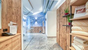A hotel reception area with a wooden reception desk, a telephone, and a glass partition. There are shelves with towels and a potted plant.