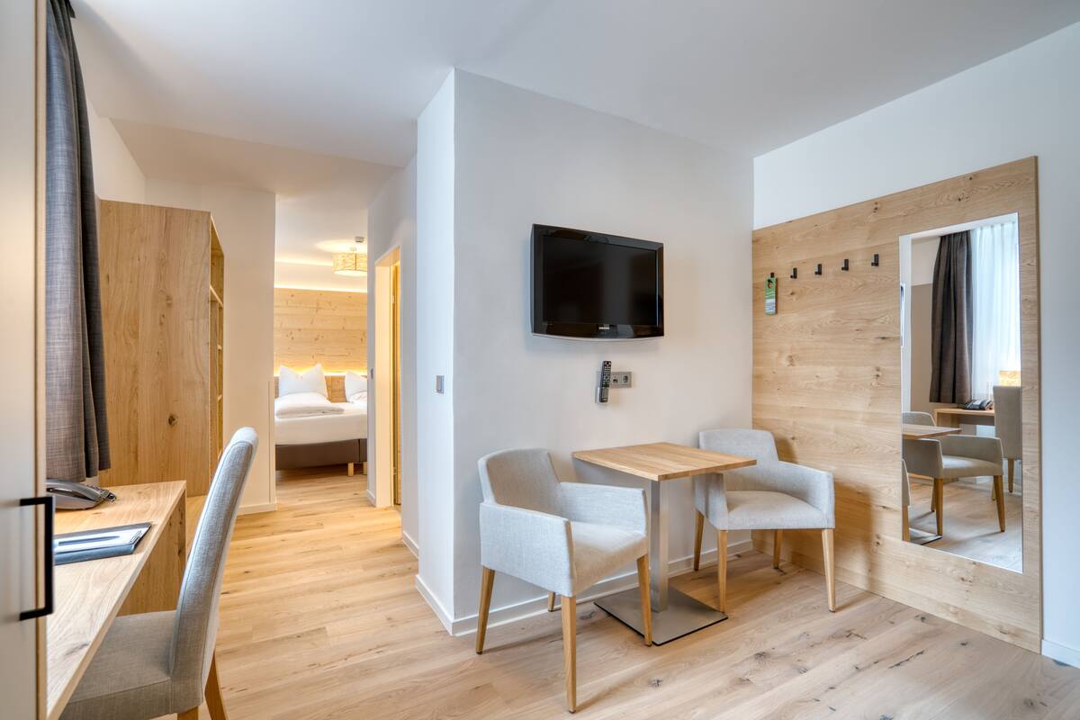 A modern hotel room with a wooden floor, a bed, a desk with a chair, a small table, and a TV mounted on the wall.