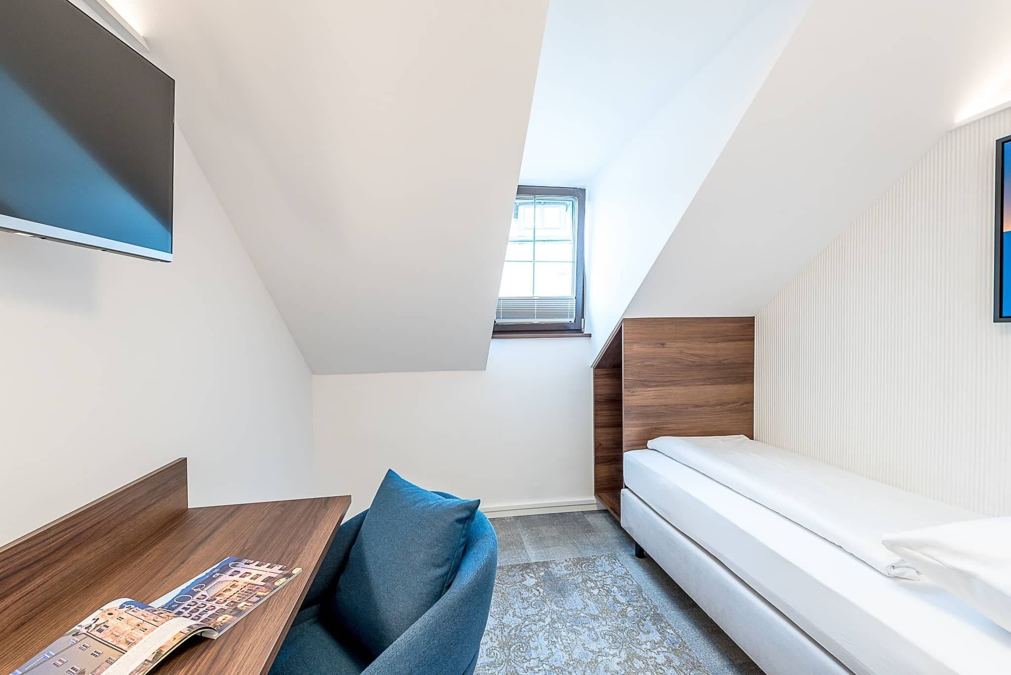 Ein kleiner, moderner Raum mit schrägem Dach, einem Bett, einem Schreibtisch mit einer Zeitschrift und einem Fenster mit Tageslicht.