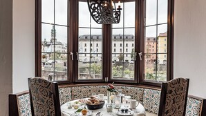 A dining table set with plates, glasses, and a vase with flowers. The table is in a room with patterned upholstered chairs and a large window with a view of buildings. - Hotel Mondschein (Innsbruck)