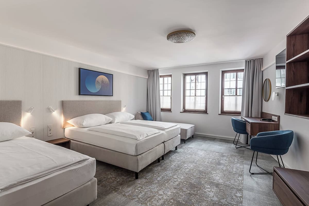A hotel room with two beds, a desk, and a chair. There are windows with curtains, a wall-mounted TV, and a ceiling light fixture.
