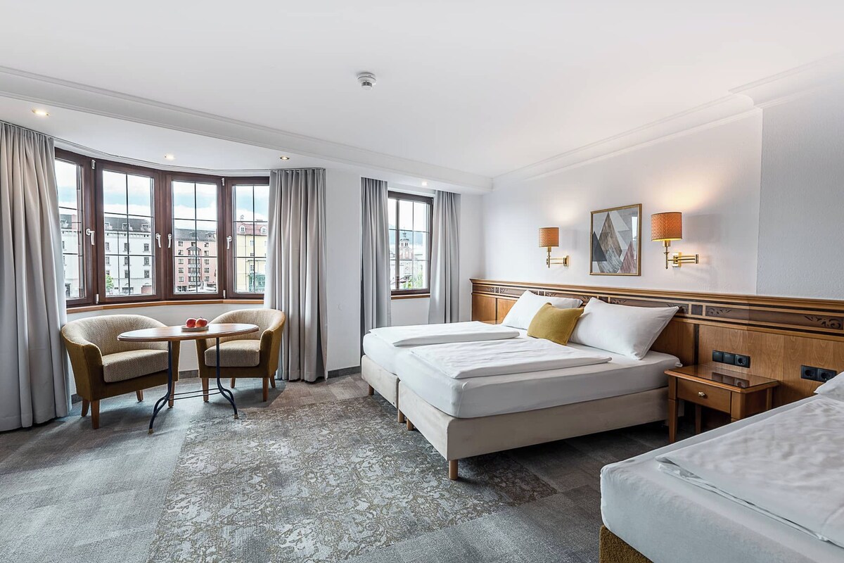 A hotel room with a large bed, a smaller bed, a sofa, a table with two chairs, and a view of the city through large windows.