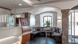 A reception area with a glass counter, a wooden reception desk, a seating area with a round table, and a stone pillar. - Hotel Mondschein (Innsbruck)