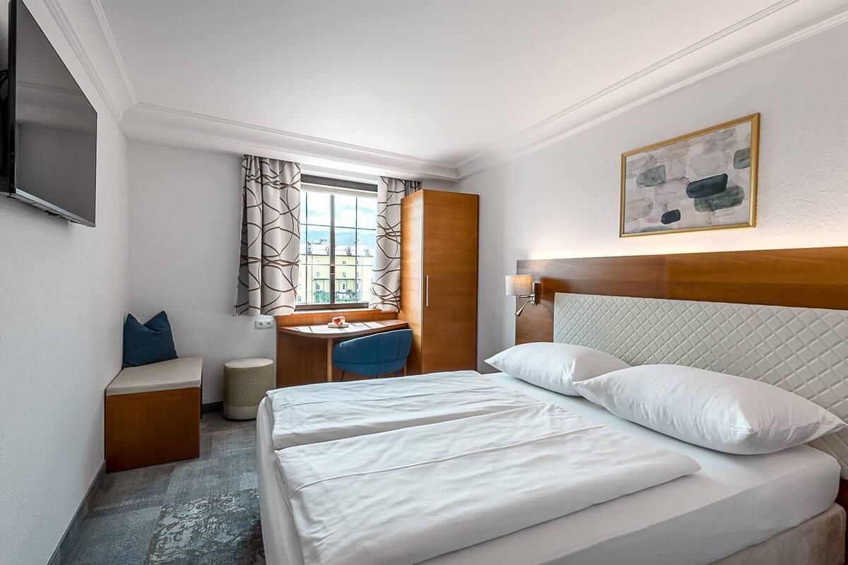 A hotel room with a bed, a desk, a chair, a TV, and a window with curtains.
