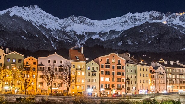 A row of colorful buildings along a river with snow-capped mountains in the background. - Hotel Mondschein (Innsbruck)