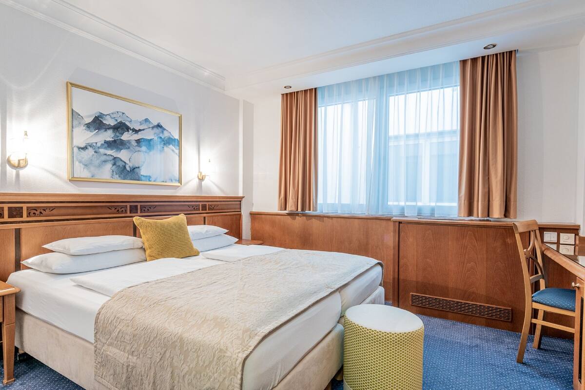 A hotel room with a large bed, a desk, a chair, a window with curtains, a wall-mounted lamp, and a painting.