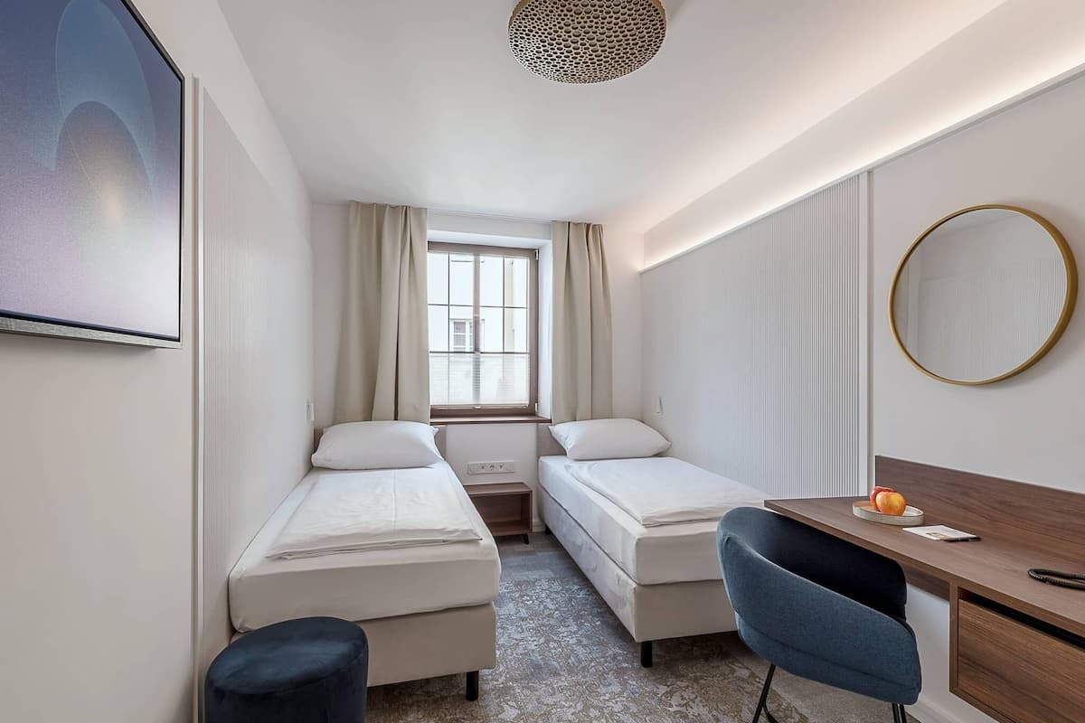 A small hotel room with two single beds, a desk, a chair, a round mirror, and a large window with curtains.