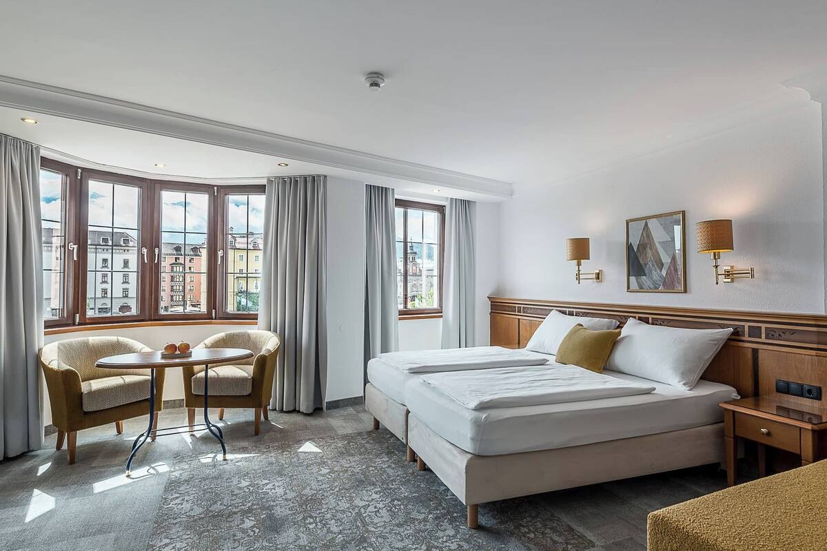 A hotel room with a large bed, two armchairs, a small table, and a view of buildings through large windows.