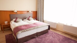 A neatly made bed with a headboard, a bedside table with a lamp, and a window with curtains. - Pannonia Hotel (Sopron)