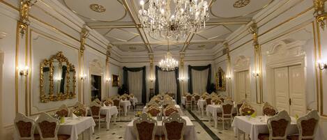 A grand banquet hall with ornate chandeliers, gold-framed mirrors, and elegantly set tables.