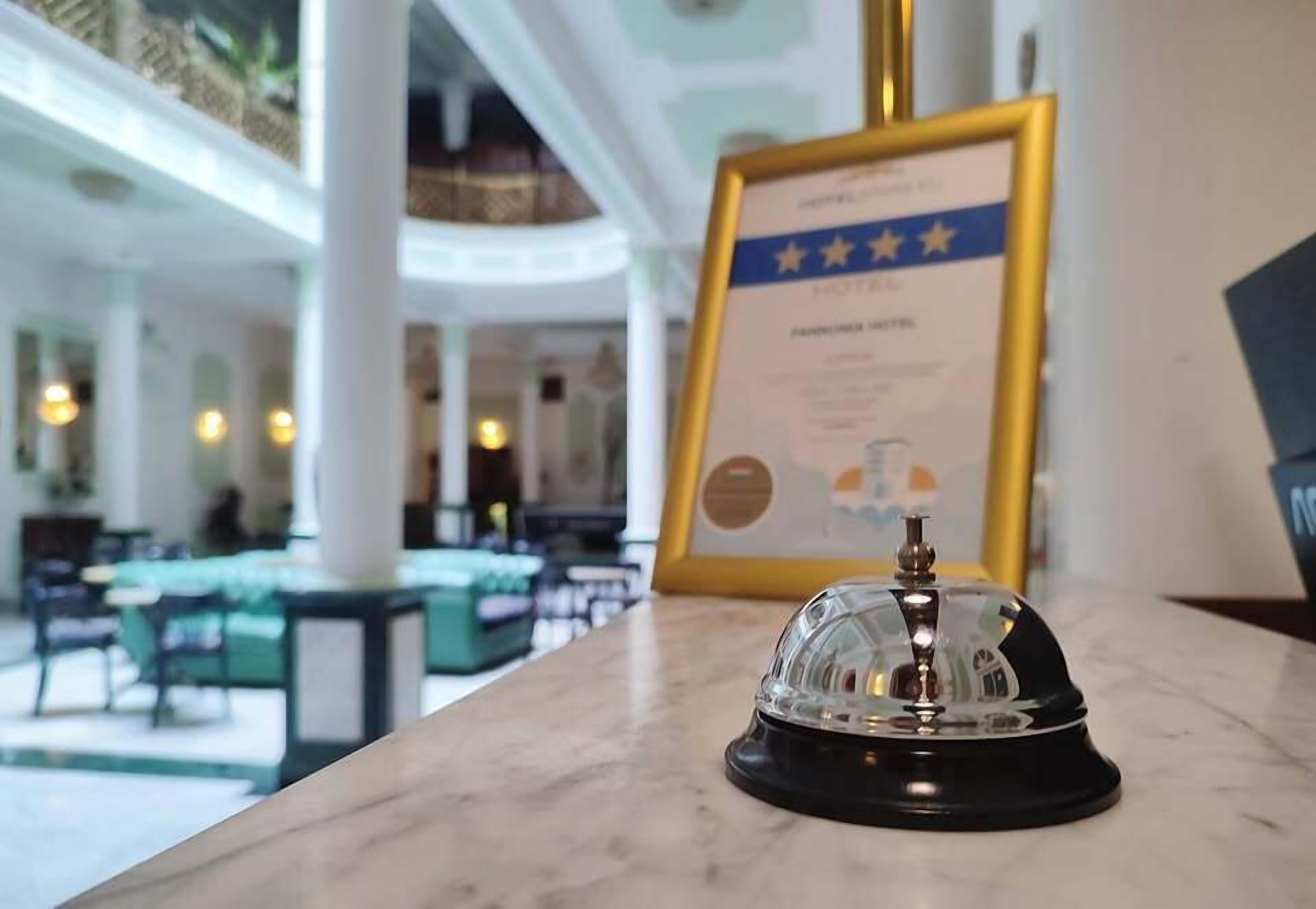 A hotel reception area with a framed award certificate on the wall, a bell on a marble counter, and a glimpse of the lobby with tables and chairs.