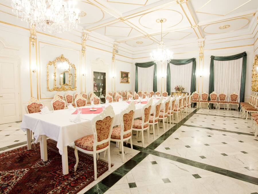 Uma sala de jantar formal com uma longa mesa posta para um banquete, rodeada por cadeiras ornamentadas, lustres e um tapete estampado.