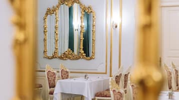 Un comedor con una mesa dispuesta para cuatro, sillas ornamentadas, una araña y un espejo grande con marco dorado.