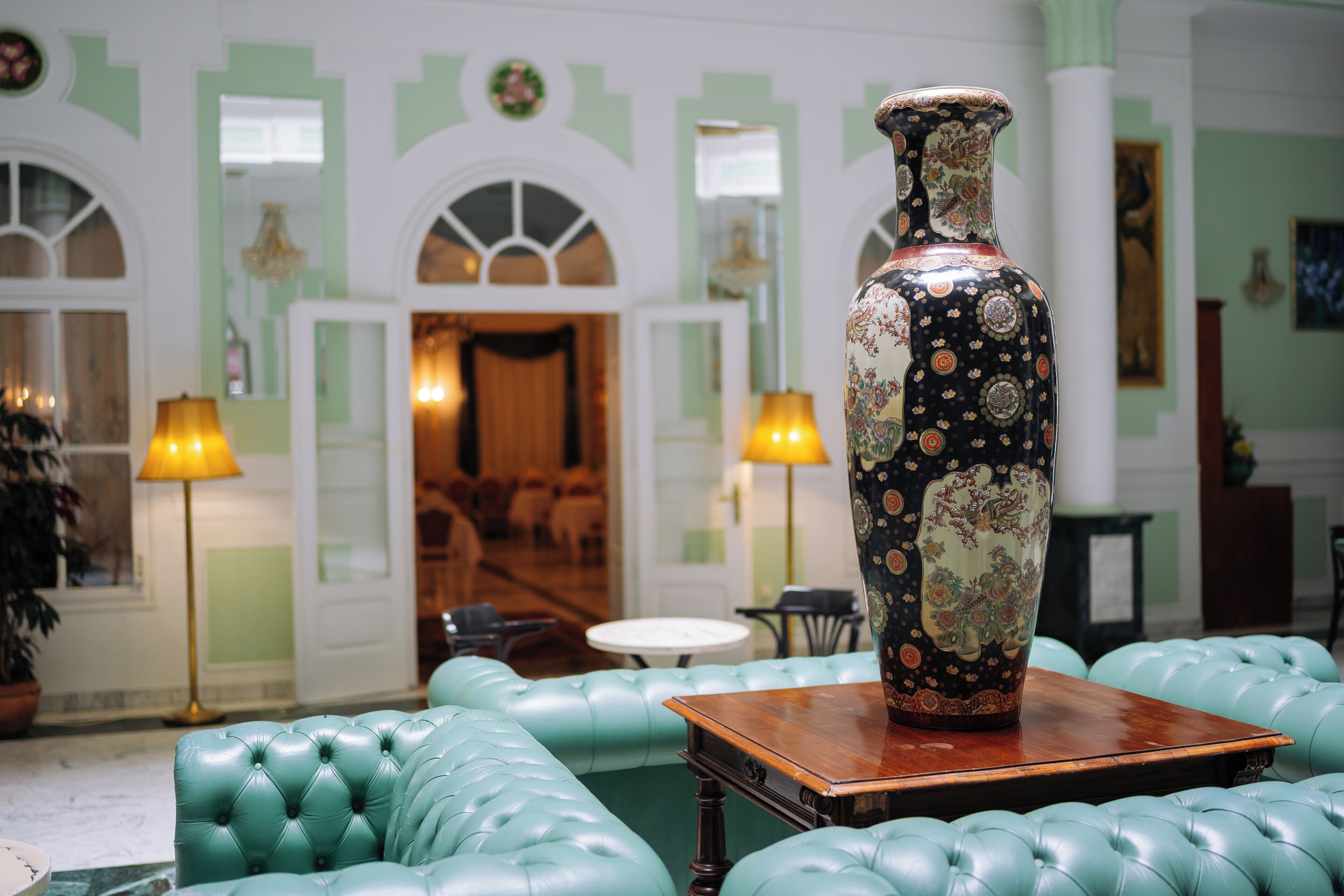 A lobby with a large decorative vase on a table, green upholstered seating, and a chandelier.
