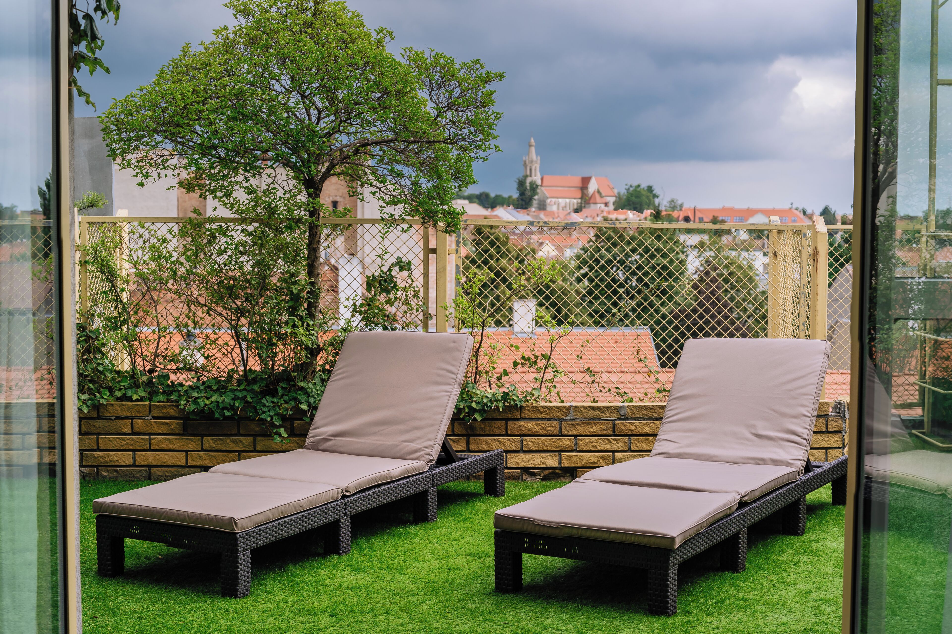 Two lounge chairs with cushions on a grassy area, a tree, and a brick wall with a fence in the background.