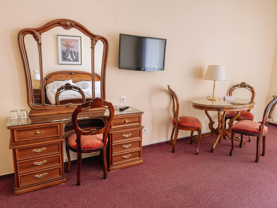 A hotel room with a wooden bed, a dresser with drawers, a vanity with a mirror, a television mounted on the wall, a round dining table with chairs, and a lamp.