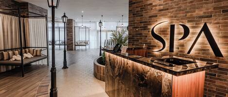 Ein Spa-Empfangsbereich mit Backsteinwand, einer Rezeption, einer Hängeleuchte und einer Sitzecke mit Bank und Kissen.