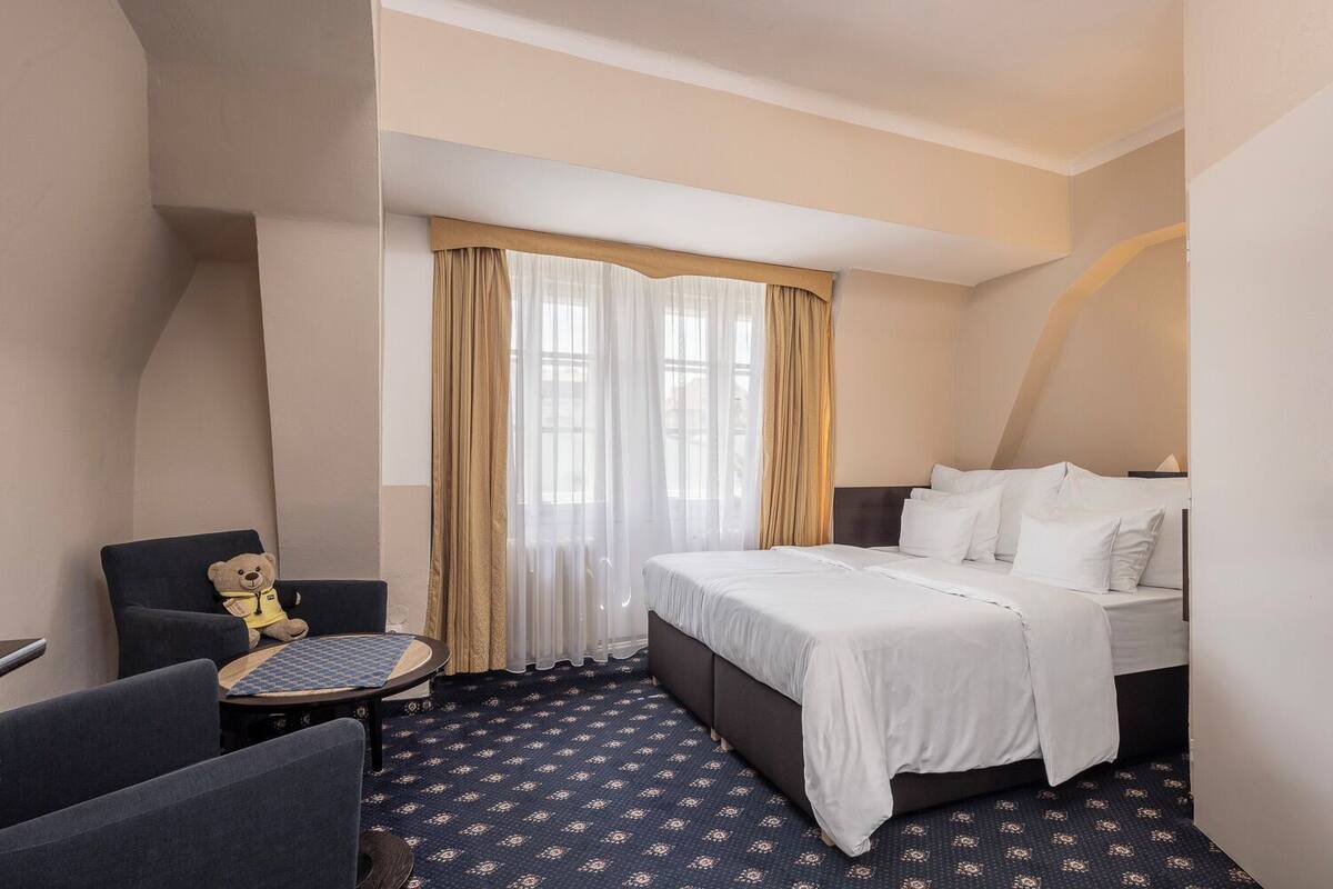 A hotel room with a bed, a chair, a small table, and a teddy bear.