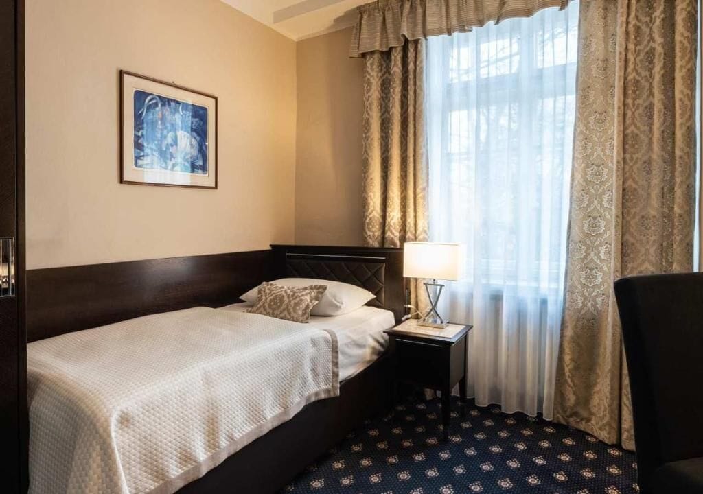 A hotel room with a bed, a nightstand, a lamp, a chair, and a window with curtains.