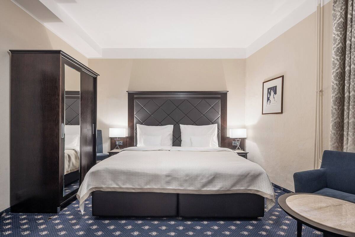 A hotel room with a large bed, a dark wooden wardrobe, a blue armchair, a small round table, and a framed picture on the wall.