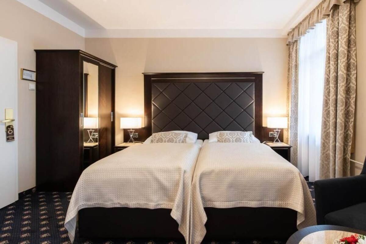 A hotel room with a large bed, two bedside tables, a dark headboard, and a large window with curtains.