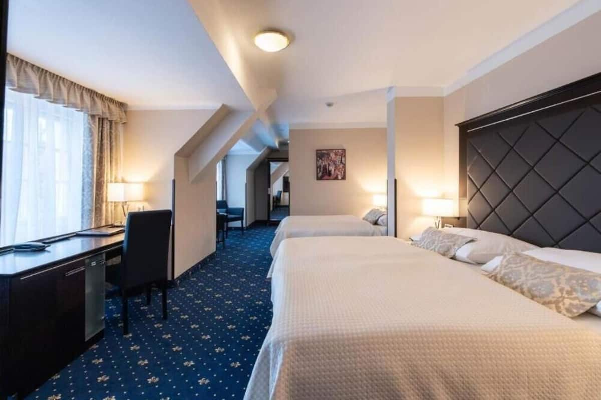 A hotel room with two beds, a desk, and a chair. There is a window with curtains, a painting on the wall, and a ceiling light.