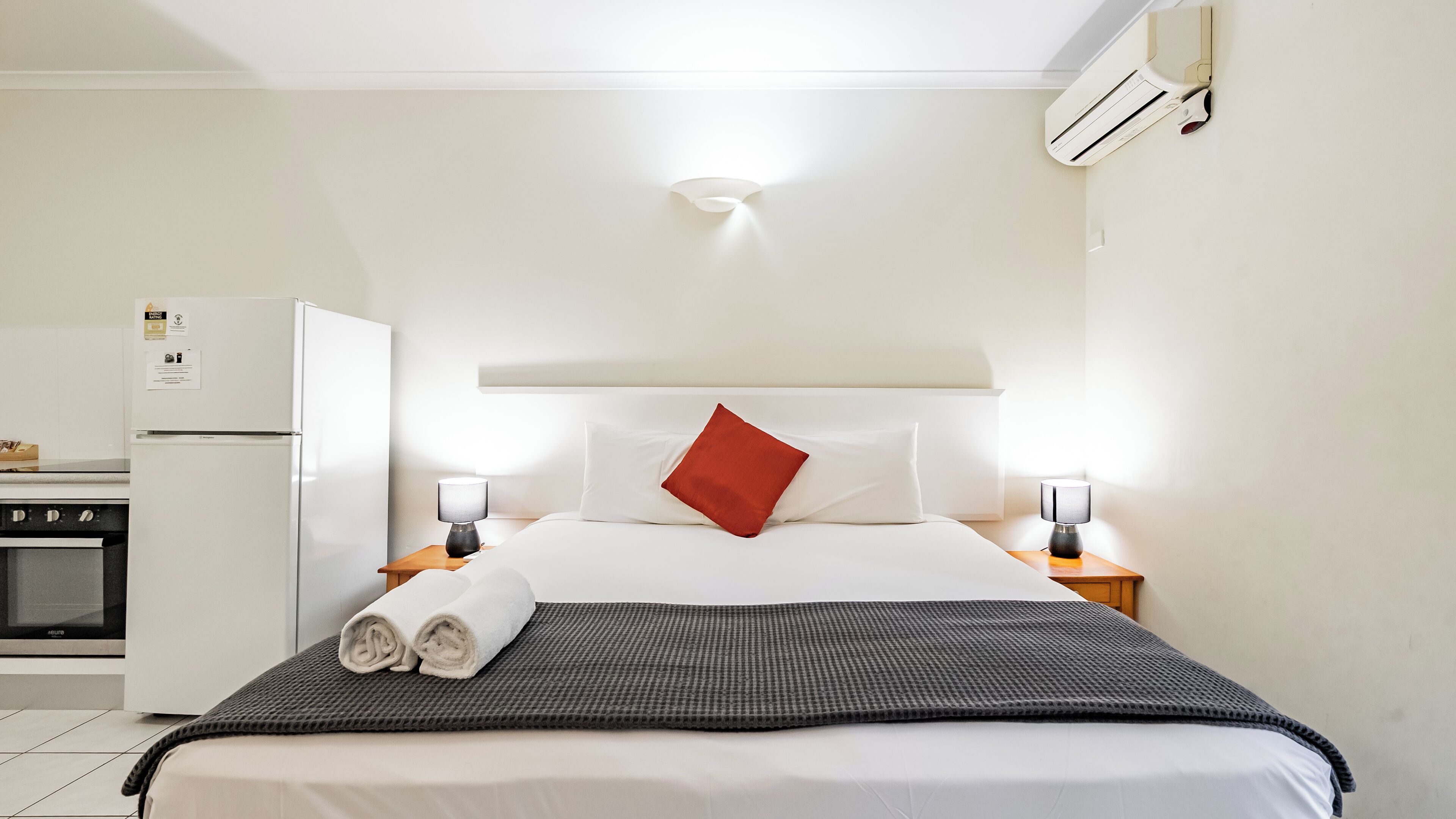 A hotel room with a bed, two bedside lamps, a red pillow, a folded towel, a small table, a refrigerator, and a microwave.