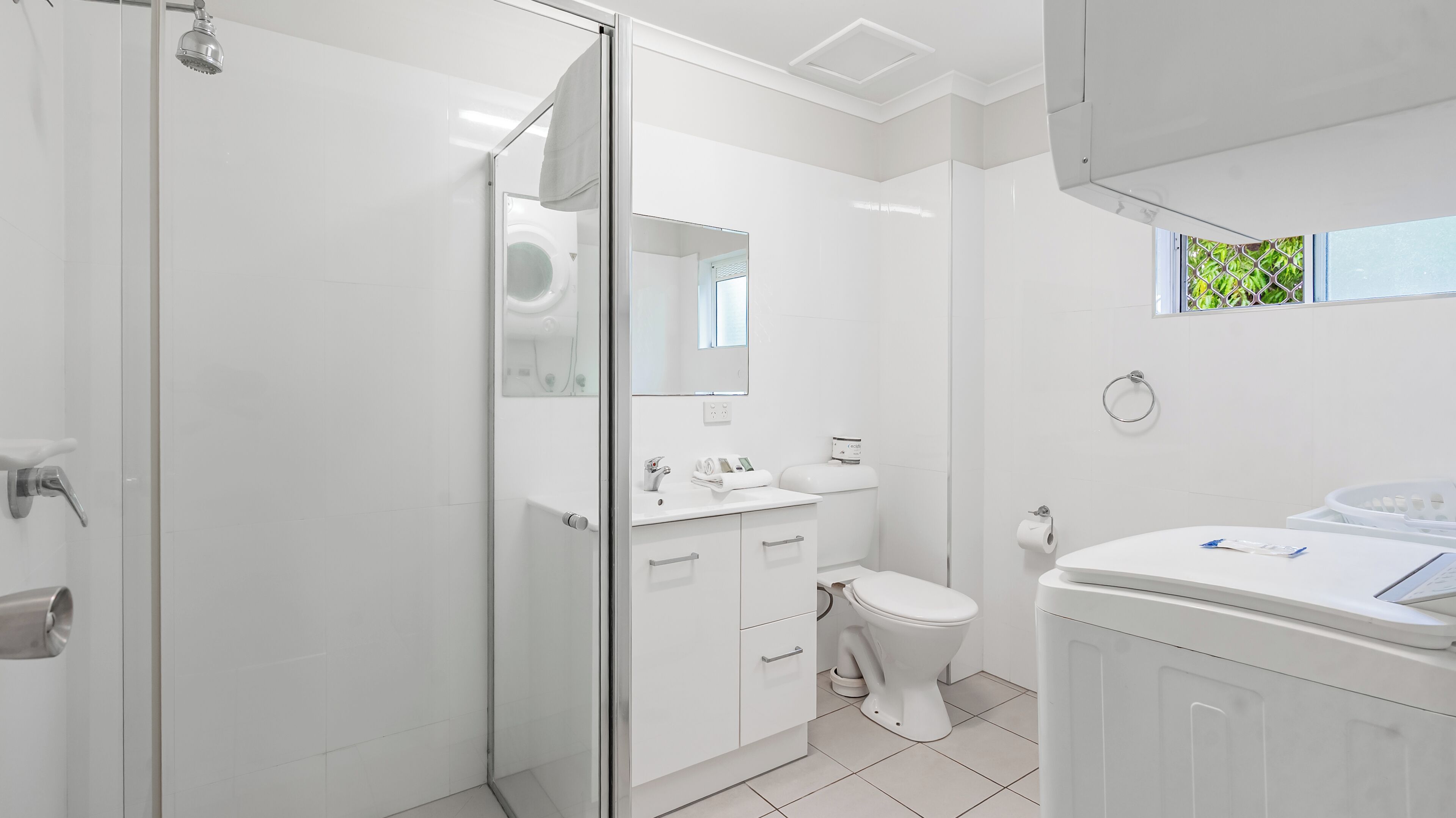 Une salle de bain moderne avec une douche à l’italienne en verre, des toilettes, un lavabo avec un miroir et une fenêtre donnant sur un espace verdoyant.