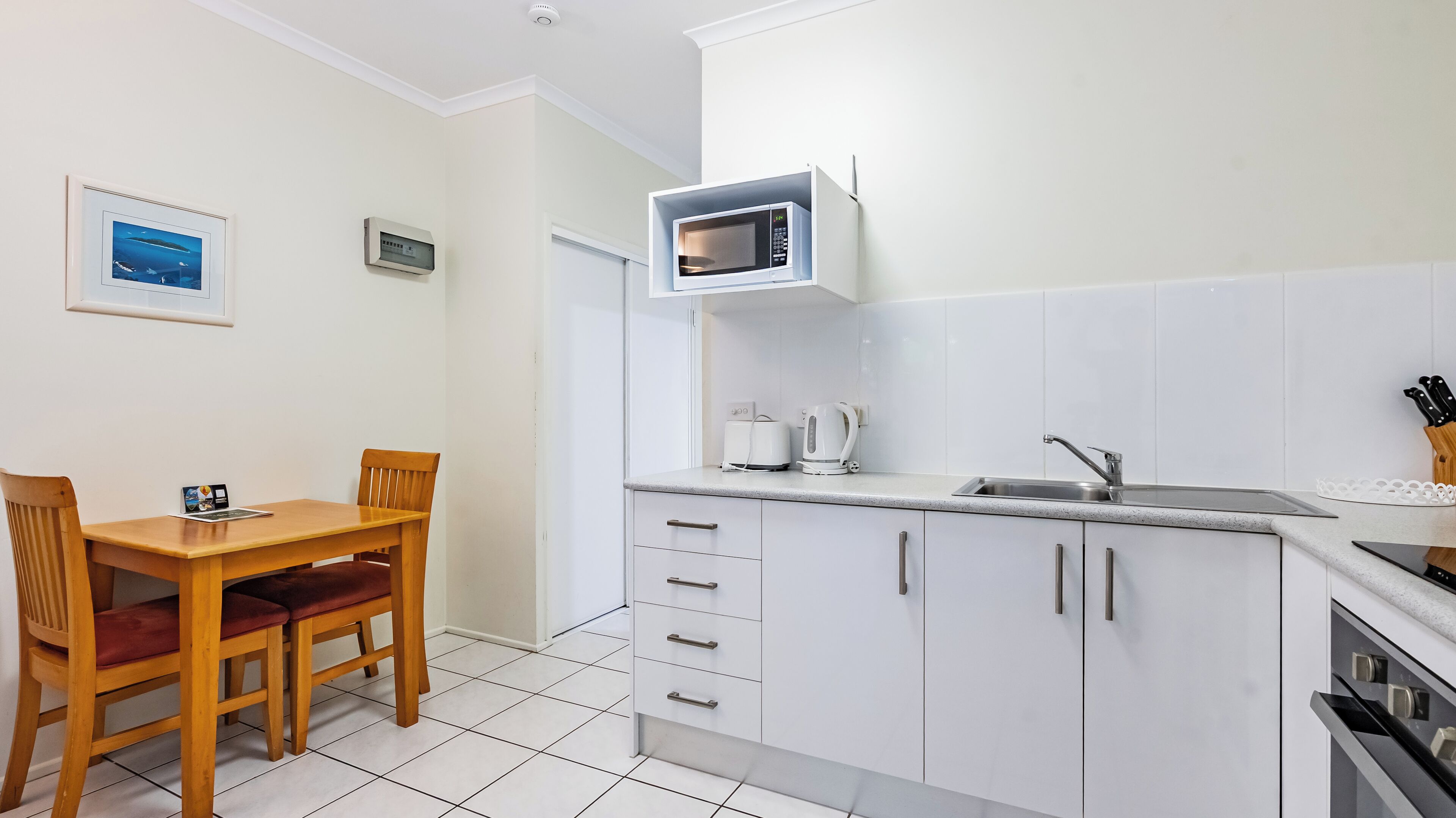 A compact kitchen with white cabinets, a microwave, a kettle, and a sink. There is a wooden dining table with two chairs.
