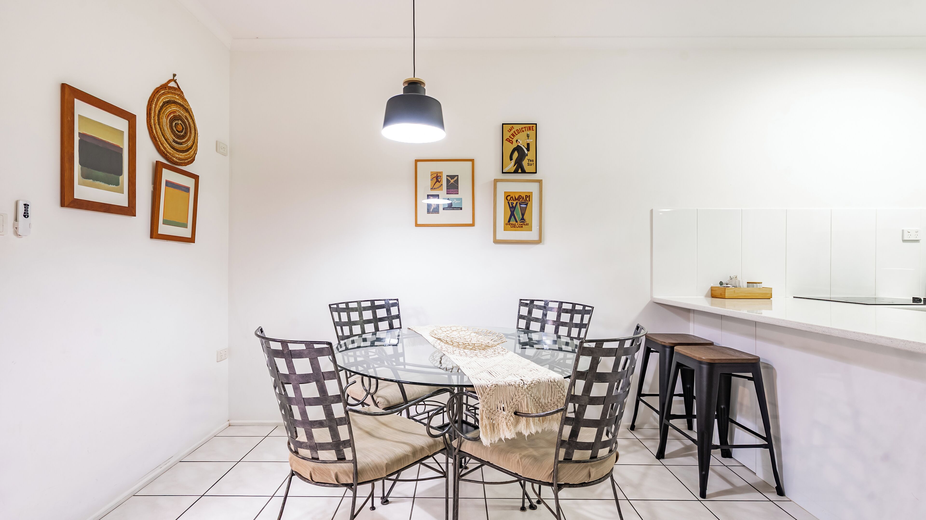 A dining area with a glass table, four chairs, a hanging pendant light, and framed artwork on the walls.