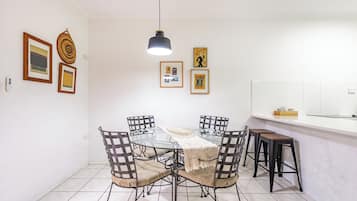 A dining area with a glass table, four chairs, a hanging pendant light, and framed artwork on the walls.
