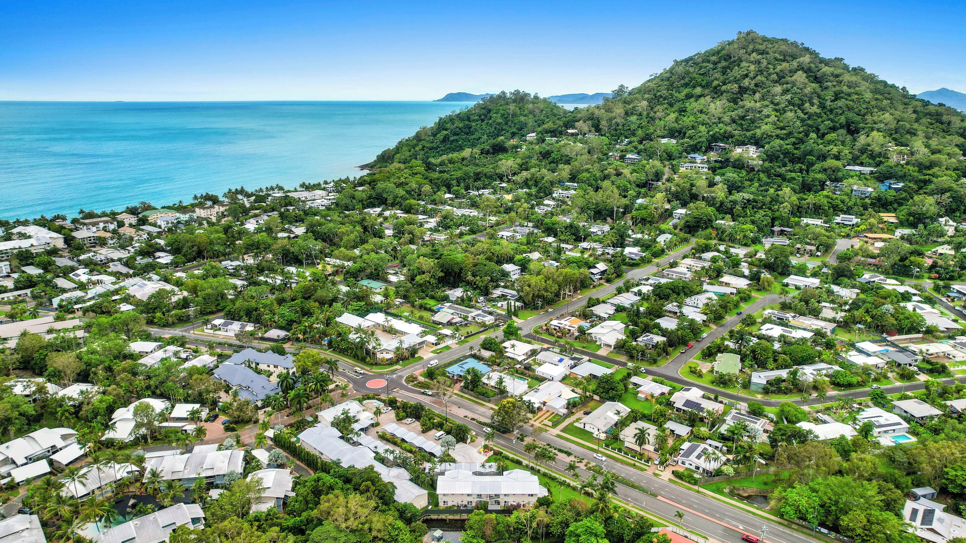 An aerial view of a coastal residential area with houses, roads, and a hill with dense vegetation.