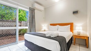 A hotel room with a bed, a nightstand, a lamp, and a view of greenery through a window. - Trinity Beach Pacific (Trinity Beach)