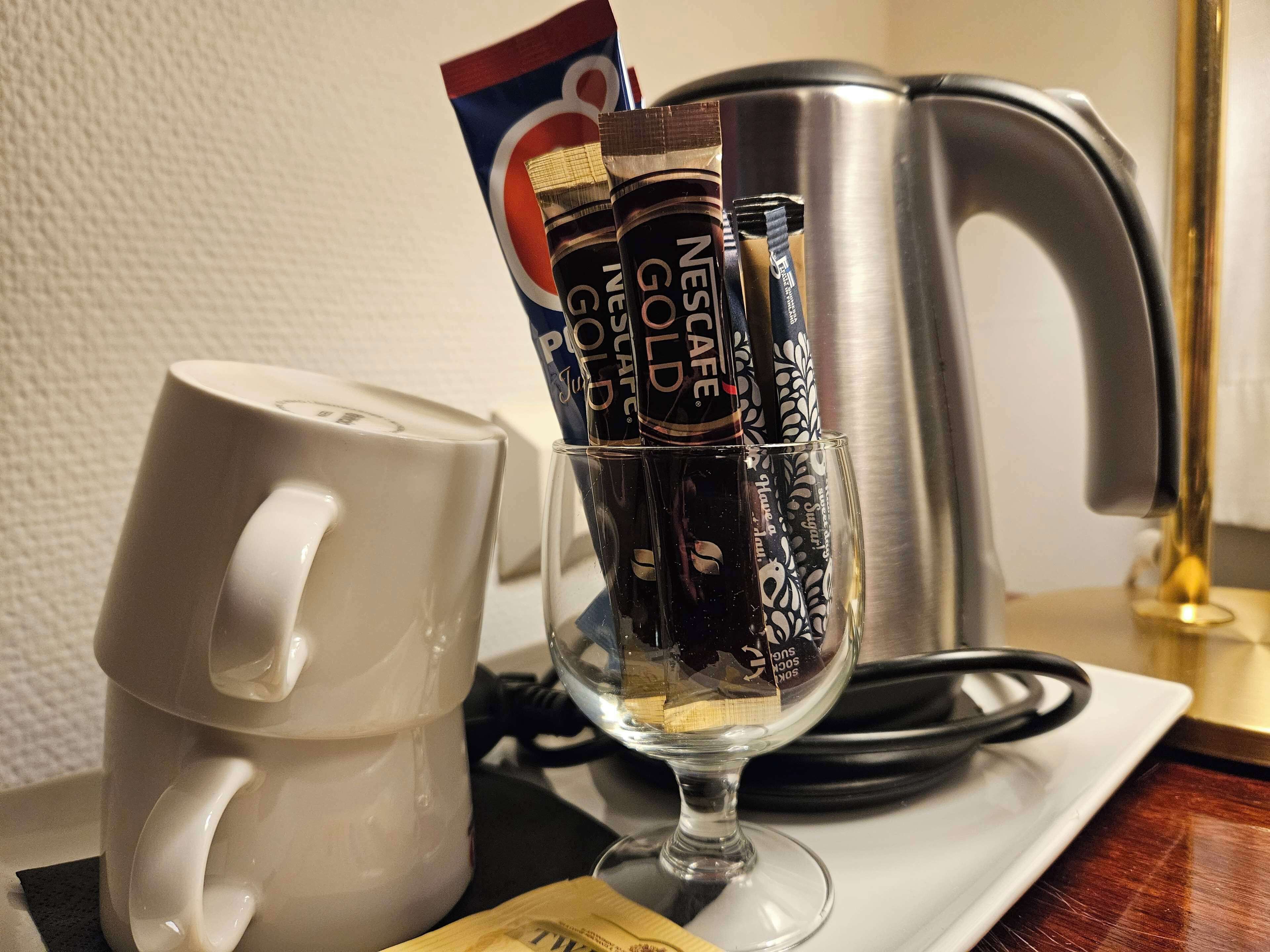 A white mug, a glass with coffee packets, and a stainless steel kettle on a table.
